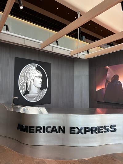 The image shows an interior view of an American Express office, likely a reception or lounge area, in Stockholm-Arlanda, Sweden. A large, circular American Express logo featuring a Roman soldier's profile is mounted on a dark gray, textured wall. In front of this logo is a curved, metallic reception desk with the words "AMERICAN EXPRESS" embossed in black, three-dimensional letters on its front surface. A sleek, modern faucet is visible above the desk. To the right of the American Express logo, a large, illuminated digital screen displays an abstract image with warm lighting and a silhouetted figure. The ceiling features exposed wooden beams and track lighting. The flooring appears to be a polished, reddish-brown material. There are no people visible in the image.