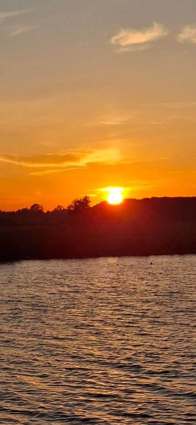 The image depicts a sunset over a body of water, likely a river or a bay, in Pärnu linn, Estonia. The sun, a bright orange orb, is partially obscured by the silhouette of a tree line along the horizon. The sky is a gradient of warm colors, ranging from deep orange near the horizon to lighter yellow and soft orange hues higher up, with wispy clouds scattered throughout. The water in the foreground reflects the colors of the sky, creating shimmering patterns on its surface. The overall scene is serene and captures the tranquil beauty of a sunset in Pärnu linn.