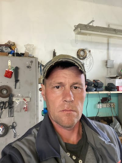 A man in a grey jacket and cap stands indoors in front of a grey metal cabinet. The cabinet contains a knife, scissors, a grinder wheel, safety glasses, and other assorted tools. Electrical outlets, tool batteries, and wires are visible on the wall above the cabinet. A fluorescent light fixture is mounted on the opposite wall. The man appears to be looking directly at the camera. The location is unspecified within the city of Foki, Russia.