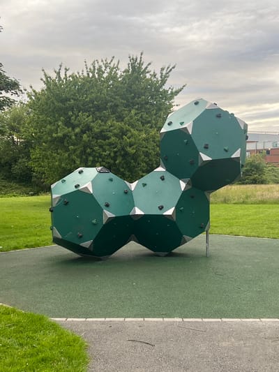 A cluster of geometric, dark green climbing structures, resembling connected octagons, is situated on a bright green rubberized play surface in a park. The structures are adorned with numerous black and some green climbing holds, as well as silver metal corner brackets. The play area is bordered by a light gray paved path and a strip of grass. In the background, a dense green tree line separates the park from a light-colored building with red accents and windows, indicative of an industrial or commercial establishment. The sky above is overcast with gray clouds. This scene is located in Balbriggan, Ireland.