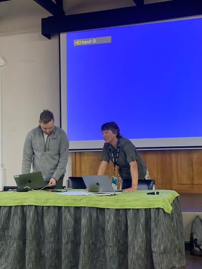 The image captures an indoor setting, likely a meeting or presentation room, in Riara Ridge, Kenya, featuring two men engaged with technology. A large projection screen dominates the left side of the frame, displaying a solid blue background with a white "Input-D" message and an 'X' symbol in the bottom left, indicating that a video input is not currently connected.
In the center, two men stand behind a table covered with a light green tablecloth and a darker green pleated skirt. The man on the left wears a grey quarter-zip sweater and is leaning over a dark-colored laptop, looking intently at its screen. The man on the right wears a dark green long-sleeved shirt and has a lanyard around his neck, bearing a yellow-bordered badge with faintly visible "NATIONAL GEOGRAPHIC" text. He is leaning over the table, seemingly interacting with a silver laptop or other items. Both laptops have the visible Apple logo.
A microphone and some documents are also present on the table. The background includes light-colored walls and wooden paneling. A black structural beam is visible in the upper left of the image. The scene suggests a professional environment, with the men focused on setting up or managing equipment for a presentation or event. The time of day is not discernible, but the space is well-lit.