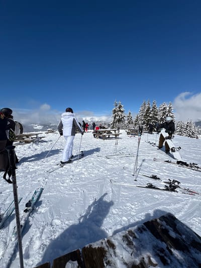 The image depicts a high-altitude plateau or summit within the ski resort of La Clusaz, France, on a clear, sunny winter day. The ground is entirely covered in granular snow, showing tracks from skis and footsteps. The sky is a deep, cloudless blue overhead, with some white, scattered clouds visible along the distant horizon.

In the foreground, leaning against an unseen object on the left, is a pair of grey ski poles with blue accents. Below them, two pairs of skis lie flat on the snow. In the lower right, the snow-dusted edge of a rough wooden structure, possibly a table or bench, is visible.

Multiple individuals, dressed in winter ski attire, are present. In the mid-ground, slightly left of center, a person wearing a white ski jacket with grey shoulders, white ski pants, and a dark helmet stands on skis, holding ski poles, facing away from the camera towards the distant landscape. To their right, a person in a dark jacket and helmet holds a white snowboard vertically. Several other pairs of skis and poles are resting on the snow between these individuals.

Further into the mid-ground and background, multiple wooden picnic tables with benches are distributed across the snow, some partially covered. Several individuals are visible around these tables; some appear to be seated or standing, dressed in various colored ski jackets, including red and dark tones. Behind the picnic tables, a dense cluster of snow-laden evergreen trees forms a natural windbreak or landscape feature. In the far background, snow-capped mountain peaks and a valley are discernible under the clear sky. Long shadows of people and objects are cast upon the snow, indicating bright sunlight.