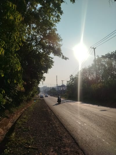 The image displays a sunny daytime street scene in Đăk Xú, Vietnam. A wide asphalt road, marked with faint dashed white lines, recedes into the distance. A single individual on a dark motorcycle travels away from the camera in the right lane. Several light-colored cars are also visible in the distance, moving in the same direction on the left side of the road. The sun, low in the sky and located in the upper right quadrant, casts a bright lens flare across the image and illuminates the road surface, creating noticeable shadows.
To the left, dense green foliage and trees line a dirt and sparse grass verge. To the right, utility poles supporting power lines run parallel to the road, which is bordered by a strip of dark, coarse aggregate material adjacent to the asphalt, followed by more trees and undergrowth. In the background, a tall cellular communication tower and multiple urban buildings are visible, set against a backdrop of distant hills or mountains under a clear, pale blue sky.