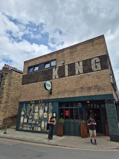 The image shows the exterior of King Street Brewhouse, a pub located at 56 King Street, Cambridge, United Kingdom. The building is constructed of light-colored brick with a large, faded "KING" logo painted on the upper facade. Below the logo, there is a mural depicting brewing equipment and wheat stalks. A circular sign identifies the establishment as "KING STREET BREWHOUSE". Two individuals are present on the sidewalk in front of the building. One, a woman wearing dark trousers and a dark tank top, is standing near the mural, looking at her phone. The other, a man in khaki shorts and a dark polo shirt, is standing by the entrance, also looking at his phone. The building features dark green painted doors and windows, and a dark green tiled section on the corner. The sky is overcast with scattered clouds. The scene is captured from a street-level perspective, showing the pavement and a road with yellow lines in the foreground.