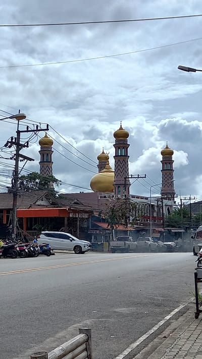 The image shows a mosque in Ban Ao Nang, Thailand. The mosque has a large golden dome and two minarets. The mosque is located on a street with power lines and buildings on either side. There are cars and motorcycles parked along the street. The sky is cloudy.