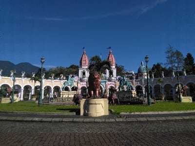 The image displays a wide view of a grand, ornate temple-like complex located in Batulawang, Indonesia, under a clear, bright blue sky. In the immediate foreground, a cobblestone-patterned paved area transitions to a well-maintained green grass median. A prominent dark green, multi-lamp street lamp stands to the left of the center, with another visible further left.
The midground features an elaborate structure characterized by a long, cream-colored arcaded facade, adorned with decorative elements and smaller statues on pedestals within the arches. A large, bronze-colored lion statue on a light-colored pedestal stands centrally in front of this structure. Flanking a central entrance within the arcades are two teal-green equestrian statues on pedestals. Additional smaller, dark lion statues are positioned around the bases of these central elements.
In the background, the main building of the complex rises, featuring two prominent pink-roofed spires topped with dark flags, indicative of Hindu temple architecture. To the right of these spires, a light blue dome structure is visible. Lush green trees and dense foliage frame and surround the architectural elements. The far left background reveals dark green mountains under the bright sky, completing the scenic environment. No people are visible in the scene.