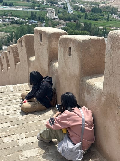 Two individuals are seated on a brick walkway along a fortified wall, overlooking a landscape in Jiayuguan, China. The person on the left wears a black jacket and tan pants, holding a red object. They are leaning against a brown backpack. The person on the right is wearing a pink hooded sweatshirt and green pants, looking at a smartphone. A grey bag with the word "Champion" is slung over their shoulder. The wall is constructed of light-colored, textured material with rectangular openings. The background shows green fields, trees, buildings, and a road with vehicles. The day appears sunny with distinct shadows.