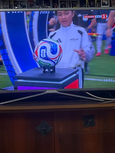 A vertical, eye-level shot captures a television screen displaying a soccer match. In the foreground, a man in a white tracksuit with grey stripes on the sleeves and black trim is holding a soccer ball. The ball is predominantly white with blue, red, and black hexagonal patterns, and features the FIFA logo and what appears to be a star design. The man is pointing at the ball with his left hand.
Behind the man, a scoreboard graphic is visible on the left side of the screen, displaying "НА ЖИВО" (Live) in red text with white lettering and a white square with "1" inside. To the right of this, a time stamp reads "19:52". The background on the screen shows a green soccer field with white lines.
The television itself is a dark color with a wide stand. To the right of the TV, a wooden cabinet with a rich, dark brown finish is visible, featuring two decorative, dark square insets. The lighting on the screen is bright, suggesting an indoor setting, possibly in Cheshnegirovo, Bulgaria, as indicated. The overall mood is neutral, focusing on the sporting event and the man presenting the ball.
