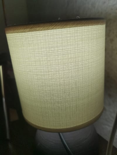 The scene, identified as being in Velichkovo, Bulgaria, depicts an indoor environment focused on a singular object. The central subject is an illuminated cylindrical lamp shade, fabricated from a light-toned, off-white or beige woven textile material exhibiting a consistent grid pattern. The upper and lower edges of the shade are bordered by a darker, possibly brown or golden-hued, trim. Below the shade, a segment of the lamp's dark, textured base is visible, from which a dark electrical cord descends, extending out of frame towards the lower left. The background is largely out of focus, comprising indistinct dark elements on the left, including a partially visible reflective vertical object, and a lighter, textured surface on the right. No human subjects or discernible activities are present.
