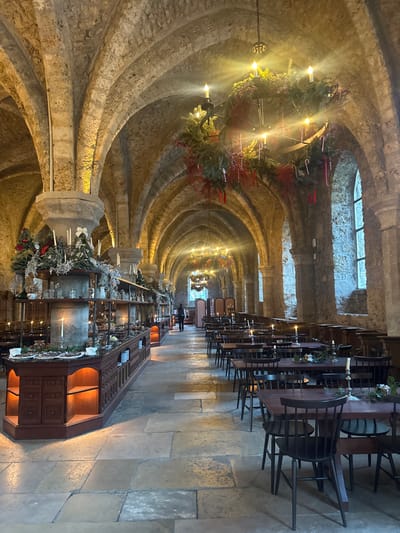 The image displays a festive setting within the vaulted stone refectory of the Abbaye des Vaux de Cernay in Cernay-la-Ville, France. The hall features high arched ceilings supported by thick stone columns and rough-hewn stone walls, with a floor composed of large, irregularly shaped stone slabs.
A long, multi-tiered dark wooden serving station extends along the left side of the hall, featuring integrated warm yellow lighting under its shelves. This station is extensively decorated with evergreen garlands, white bows, small artificial Christmas trees, and multiple white pillar candles in silver holders, with various dishes and serving items displayed on its surfaces.
Overhead, multiple large, circular metal chandeliers hang from the ceiling, each adorned with evergreen garlands, red berries, and white candy canes, and illuminated by several candle-shaped light bulbs. On the right side of the hall, rows of dark wooden rectangular tables and matching chairs are arranged. Each table is minimally decorated with a small evergreen sprig and a white candle in a silver holder. Tall, arched windows on the right wall allow natural light into the space.
A single individual, dressed in dark clothing, is visible walking away from the camera in the central aisle, positioned towards the far end of the serving station. The overall environment is set for a festive event, likely during the Christmas season.