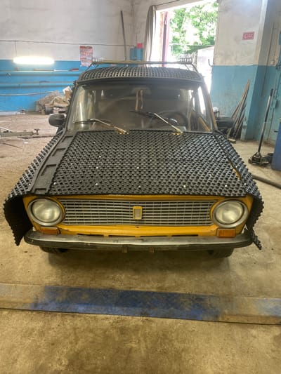 The image shows a yellow car with a unique black grille cover. The car is parked in what appears to be a garage or workshop. The grille cover is made of a textured material with a grid pattern and appears to be custom-fitted to the front of the vehicle, covering the headlights and grille.
The setting is a dusty concrete floor, with blue and light-colored walls in the background. There are some tools and equipment visible, including a car jack and what looks like metal shelving. Natural light seems to be coming from an open doorway or window on the left, revealing some greenery outside.
A red and white sign with Cyrillic text is mounted on the wall to the left of the car. The text on the sign reads "НЕ БОИРЬ", which translates to "Don't be afraid".
The image is a static shot, with no activity clearly depicted, other than the car being present in the workshop. The lighting suggests it could be daytime. The overall impression is one of a utilitarian space where a vehicle might be undergoing maintenance or modification.