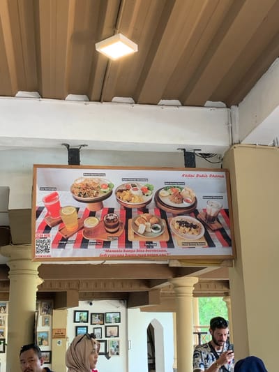 The scene is indoors at a restaurant called "Kedai Bukit Rhema" in Kecamatan Borobudur, Indonesia. The restaurant has a menu board hanging above the customers' heads, featuring photos of several Indonesian food and drink dishes. The dishes are presented in a colorful and appealing way. The menu includes "Bakmi Ekwan Goreng", "Ayam Kremes Serundeng", "Nasi Goreng Special", "Tahu Bakso", "Pisang Goreng Special", "Jus Semangka", "Orange Squash", "Lemon Tea", "Kopi Cita", and "Hazelnut Chocolate". There is a white ceiling with a single light fixture. The restaurant is located inside a building with wood and white walls. There are four pillars in the foreground, and a window with a view of green vegetation outside. In the foreground, a man in a white and blue shirt is looking at his phone. He has black hair and a beard. A woman in a white hijab is standing to his left. A second man is sitting with his back turned to the camera. There are several framed pictures on the wall behind the patrons. The image was captured in the afternoon.