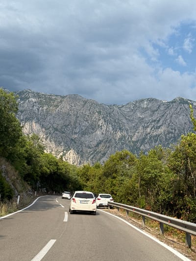 The image captures a winding asphalt road in Dražin Vrt, Montenegro, with a mountainous backdrop under a cloudy sky. Three white cars are visible on the road, two in the foreground and one further back, all traveling in the same direction. The car closest to the viewer is a Toyota Yaris, with its license plate reading "RO AT391". The road is bordered by a metal guardrail on the right and lush green trees and vegetation on both sides. The imposing, rocky mountains in the background dominate the upper half of the frame, with their rugged texture clearly discernible. The lighting suggests it is daytime, possibly late afternoon, given the shadows and the mix of sunlight and cloud cover.
