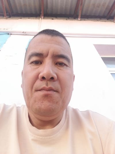 The image is a close-up selfie of a man's face and upper body, taken outdoors. The man has dark hair, dark eyes, and is wearing a light-colored t-shirt. The background shows a white building wall and a corrugated metal roof supported by wooden beams. There are also some pipes visible. The lighting suggests it is daytime. The specific location is Gumbaz, Uzbekistan.