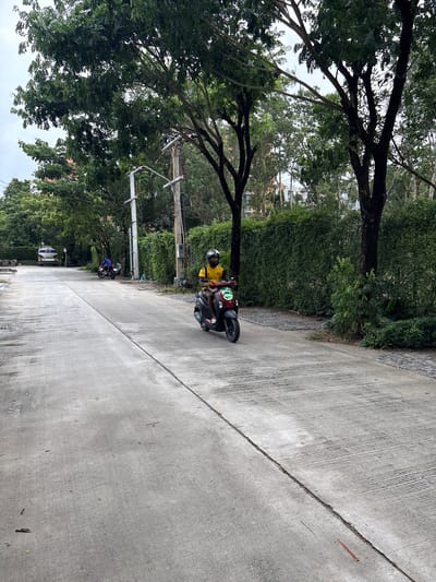 In Sakhu, Thailand, a person wearing a yellow shirt and a helmet rides a dark-colored scooter with red accents on a wet concrete road. The scooter's headlight is illuminated. The road is flanked by lush green trees and hedges, with a clear sky visible through the branches. In the distance, a white truck with a blue tank and another person on a motorcycle can be seen. The scene appears to be in a residential or semi-rural area.
