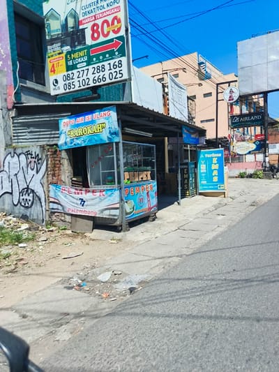The image shows a street scene in Kecamatan Margahayu, Indonesia, located at latitude -6.967 and longitude 107.576. The street is paved and has a sidewalk on the right side. There are several buildings along the street, including a two-story building with a sign advertising "CLUSTER I DEKAT TOL KOPU HARGA TERJANGKAU" with a red arrow pointing to the right and the number 800 in white letters. The building has a blue and white sign with a phone number and the words "HUB 5 MENIT KE LOKASI". There is a street vendor selling "PEMPEK PALEMBANG" with a blue and white sign and a glass display case full of food. The street vendor is located under an awning. There is a small sign on the right side of the image with the word "AIRLAND" written in white letters. The sky is blue and clear. The image is taken from the perspective of a person walking down the street.