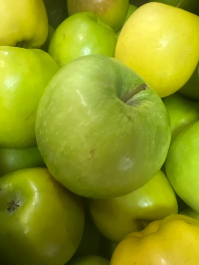 A close-up, eye-level, indoor shot shows a pile of ripe, green apples with a few yellow apples interspersed. The apples are arranged tightly together, filling the frame. The lighting is bright and natural, likely from a storefront or market. The image is devoid of human presence, traffic, or any recognizable landmarks. The absence of specific urban identifiers makes it impossible to pinpoint a location within Johannesburg, South Africa.