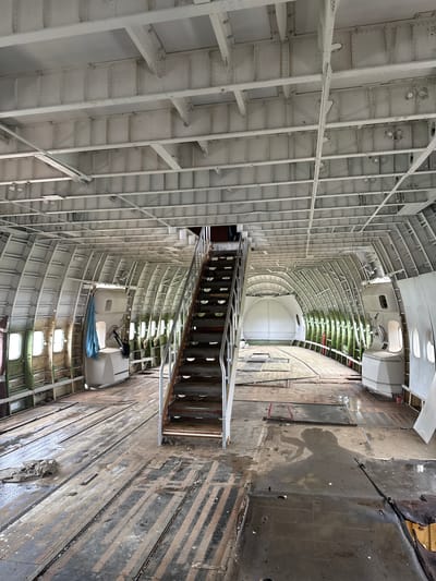 The image depicts the interior of a large, abandoned aircraft, possibly a Boeing 747, stripped down to its skeletal structure. The fuselage's characteristic curved ribs are exposed, painted a weathered white. Metal beams and crossbeams crisscross the ceiling, also painted white. The floor is made of wooden planks and metal plating, showing signs of wear, dampness, and debris.
A prominent set of wooden stairs with metal railings ascends towards the upper deck, which is not fully visible. To the left of the stairs, a white structure, likely a lavatory or service unit, is attached to the fuselage wall, with a blue cloth hanging from it. Several circular windows, framed in white, punctuate the curved walls along the length of the aircraft. In the distance, the curved front of the aircraft is visible, with a white dome-like structure. Green material, possibly insulation or remnants of seats, is visible along the lower sections of the fuselage walls.
The overall scene suggests a derelict state, with a pervasive sense of disuse and decay. The presence of water on the floor indicates potential leaks or exposure to the elements. The lighting is natural, coming from the windows and possibly openings, casting shadows that add to the somber atmosphere. The image captures an interior view of a deconstructed aircraft, emphasizing its structural framework and the passage of time. This scene is located in Pak Nam Pran, Thailand, suggesting the aircraft is being repurposed or has been grounded in this coastal district.