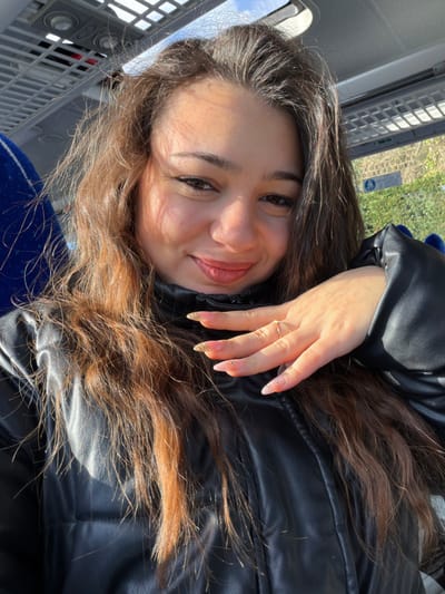 A young female, with long wavy brown hair and wearing a black puffer jacket, is depicted seated inside a passenger vehicle, likely a bus or coach. She is smiling and looking directly at the camera, with her left hand raised towards her chin. Her left hand displays almond-shaped artificial nails; the index, middle, and ring fingers feature gold glitter tips, while the thumb and pinky have a clear/light pink base with small, clear embellishments. A simple gold-colored ring is present on her left ring finger. The vehicle's interior shows a blue upholstered seat on the left and overhead racks and lighting. Through a window behind the subject, green foliage, a bright blue sky, and a partially visible French sign with the text "VOIE DE SECOURS" are discernible. The vehicle is situated in France; the precise location in the city of Bouxwiller is not discernable from the image. Natural light illuminates the subject's face, casting shadows.