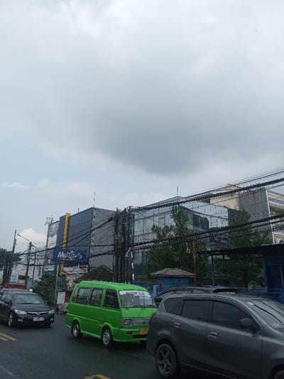 A street scene features a green van with "Sparco" on its windshield, parked between a black sedan and a gray SUV. Buildings loom in the background, including one with a blue and yellow sign. Utility lines crisscross the sky above the structures. The overcast sky is predominantly gray and white. The street appears wet, with visible lane markings.