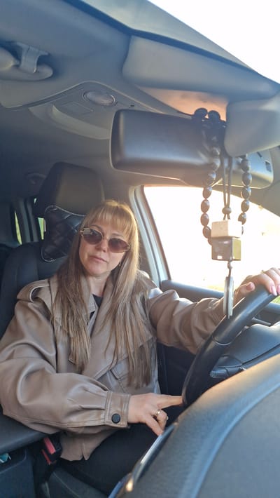 A woman with light brown hair and sunglasses sits in the driver's seat of a car. She wears a beige leather jacket and holds the steering wheel. A beaded car freshener hangs from the rearview mirror. The seat behind her has a quilted headrest. The car is in Otchor, Russia.
