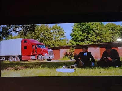 In Zilupe, Latvia, a red semi-truck with a white trailer is parked on a grassy area. Two young males are sitting on the grass in front of the truck, facing away from it. The male on the left is wearing a black suit with a white bow tie and is holding a cup. The male on the right is wearing a black long-sleeved shirt and is looking at a mobile phone. A white rectangular object is on the ground between them. In the background, there is a brick wall and green trees under a blue sky.