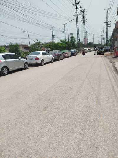 The image shows a street scene in Peshawar City Tehsil, Pakistan, on a bright, cloudy day. A line of cars is parked along the left side of the road, and more vehicles are visible further down the street, including a motorcycle with a rider and two pedestrians. Power lines and utility poles dominate the background, suggesting an urban or semi-urban environment. The road surface appears to be asphalt.