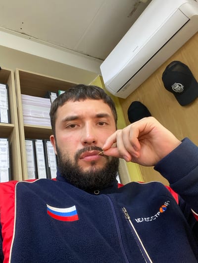 A man with a beard is sitting in an office in Luzhitsy, Russia. He is wearing a dark blue fleece jacket with red accents and a patch of the Russian flag on the left chest. He is holding a toothpick between his lips. Behind him, shelves are filled with binders. An air conditioner is mounted on the wall above him, and a black baseball cap with a skull logo is hanging on the wall to his right.