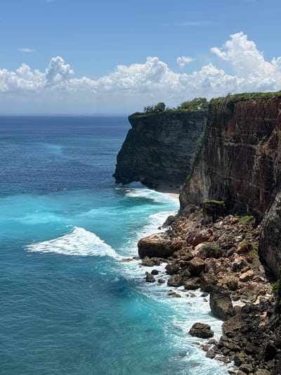 The image presents a coastal scene in Uluwatu, Indonesia, featuring a towering cliff face meeting the ocean. The cliff is comprised of layered rock formations, with vegetation clinging to its upper edge. The ocean is a vibrant turquoise, with white waves crashing against the base of the cliff and along the coastline. Cumulus clouds are visible in the blue sky above the scene. No people are visible.