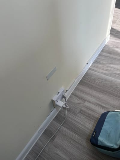 The image captures an indoor scene, focusing on the junction of a light-colored wall and a grey wood-look laminate floor. The perspective is a downward, diagonal shot.
A white power strip, equipped with at least three AC outlets and two USB ports, is mounted on the wall. Two white power adapters or chargers are plugged into the power strip, with white cables extending from them. One visible cable runs horizontally along the base of the wall.
A white cable management channel runs horizontally along the base of the wall, likely concealing wires. Above this channel, another thinner white strip, possibly also for cable management, is visible. Further to the left on the wall, a rectangular grey adhesive strip is applied.
In the upper right portion of the frame, a blue and light blue soft-sided bag is partially visible on the floor. The brand name "bozone" is discernible on the bag. The floor exhibits a distinct grey wood grain pattern. The lighting is bright and even, consistent with an indoor setting during the day. No people are visible. The scene depicts a practical arrangement for power access and cable organization.