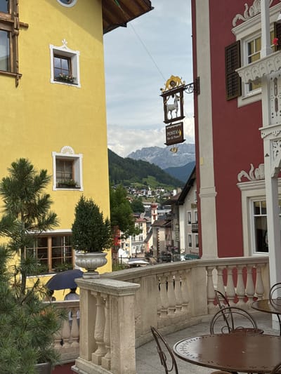 The image depicts a view of the town of Ortisei, Italy, from a balcony area. In the foreground, a balustrade with stone pillars separates the viewer from a patio with outdoor seating, including round tables and metal chairs. To the left, a vibrant yellow building is visible, featuring traditional wooden window frames and window boxes with flowers. To the right, a red building displays a decorative white facade detail above a window. A sign hanging from a decorative wrought-iron bracket on the red building reads "POSTA'S Bar & Piano" and features a depiction of a white animal, possibly a goat or sheep, above it.
Beyond the immediate buildings, the town of Ortisei unfolds with a collection of colorful houses, many with pitched roofs, nestled on a hillside. In the background, a majestic mountain range rises, its peaks partially obscured by clouds. The sky above is overcast. In the mid-ground, near the yellow building, two figures are partially visible beneath a dark umbrella, suggesting it might be raining or the sun is strong. A few evergreen trees are strategically placed, including one in a large planter, adding greenery to the scene. The overall impression is a charming alpine town with a blend of traditional architecture and natural beauty.