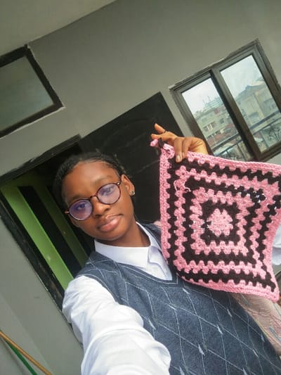 A young female subject is depicted indoors, framed against a light grey wall. She has dark hair styled in braids, wears round, dark-framed glasses, and a small stud earring in her right ear. Her attire comprises a white collared shirt and a dark grey V-neck sweater vest with a diamond pattern. Her lips are glossed. The subject holds a square-shaped crocheted item in her right hand. This item exhibits an alternating pattern of light pink and dark brown yarn, forming concentric squares. The background features a dark-framed opening to the left and a large, dark-framed window to the right. Beyond the window, an urban environment is visible, characterized by multiple concrete or stucco buildings, suggesting an elevated view within a city. The setting is identified as Cotonou, Benin.