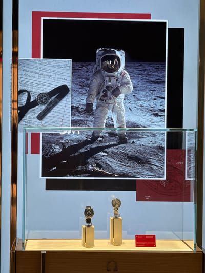 The image shows a window display of a luxury watch store, likely Omega, in Nong Prue Subdistrict, Thailand. The display features a large photograph of an astronaut on the moon, a vintage document with the Apollo mission name, and two Omega watches on stands. The astronaut photograph is prominently placed in the upper half of the display. Below the astronaut image, a document is partially visible, showing handwritten text and details about the Apollo mission and the Omega brand. The lower half of the display showcases two Omega watches, one on a gold-colored stand and another to its right. A red card with text is also visible in the lower right of the display. The entire display is framed by a light-colored border and set within a glass cabinet.