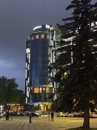 The image depicts a tall, modern building with numerous illuminated horizontal lines, likely a hotel or office complex, at night. The building has "360 RESTAURANT" illuminated on its side. Trees are partially obscuring the view of the building. Along the right side of the image is a street with parked cars and pedestrians. The location is Komgaron, Russia.