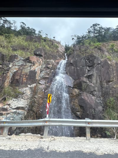 The image captures a scenic view of a waterfall cascading down a rocky mountainside, possibly along a roadside in Nam Khanh Vinh, Vietnam. The waterfall is the central focus, with water flowing over jagged brown and grey rocks. Green vegetation, including grasses and trees, grows on the slopes of the mountain, contrasting with the rocky terrain.
In the foreground, a metal guardrail runs horizontally across the frame, indicating the presence of a road. The guardrail is silver and appears sturdy, with some yellow and red reflective markers. Beyond the guardrail, a portion of the road surface is visible, appearing to be paved with asphalt and gravel.
A yellow chevron sign with a black arrow pointing left is mounted on a pole beside the waterfall, suggesting a curve in the road or a hazard. The sky above is overcast and a pale grey, hinting at cloudy weather. The overall scene depicts a natural landscape with elements of infrastructure, suggesting it's a location accessible by road. There are no people or animals visible in the image.