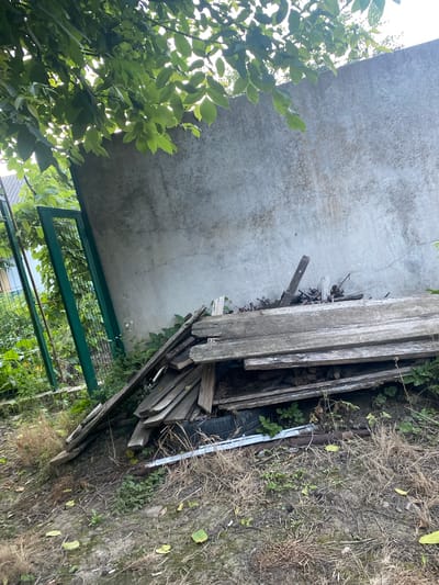 This is an outdoor, eye-level, medium-shot photograph taken on a sunny day. The image depicts a cluttered area with a concrete slab in the upper left, leaning against a pile of weathered wooden planks and debris. Green foliage, possibly from a tree or bush, hangs into the frame from the left, partially obscuring the concrete. A green metal fence runs along the bottom of the image, with a wire mesh section visible. Beyond the fence, more greenery and what appears to be a building can be seen. The ground is covered in dry grass and dirt, with some scattered green plants. The overall impression is one of an unkempt outdoor storage or yard area, possibly in a rural or semi-rural setting, given the location context of Zastavna, Ukraine. There is no text visible in the image.