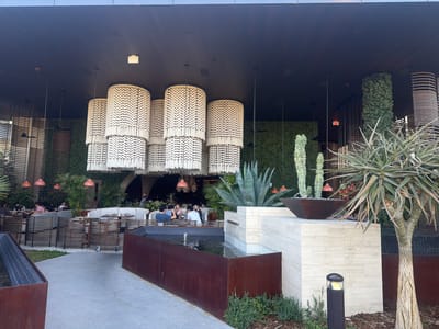 The image captures an outdoor dining area, likely in Las Vegas, United States of America. Diners are seated at tables under a covered area, illuminated by decorative cylindrical light fixtures with fringed edges. A water feature is visible in the foreground, with rectangular metal containers holding water and a tiled structure supporting various plants, including cacti and agave. A distinctive tree is positioned to the right. The architectural style incorporates a blend of modern design with natural elements like greenery and wood.