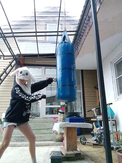 A person wearing a black hooded sweatshirt and light-colored shorts is punching a large blue punching bag that is suspended from a metal frame. The person is also wearing a white hat with bunny ears. The scene appears to be an outdoor area, possibly a backyard, with a covered patio area. In the background, there is a house with windows and siding, a staircase, and some potted plants. There are also two scooters visible in the scene, as well as a table with a blue box and some other objects on it. The ground is concrete. The lighting suggests it is daytime. The specific location in the city of Psebai, Russia, cannot be determined from the image alone.