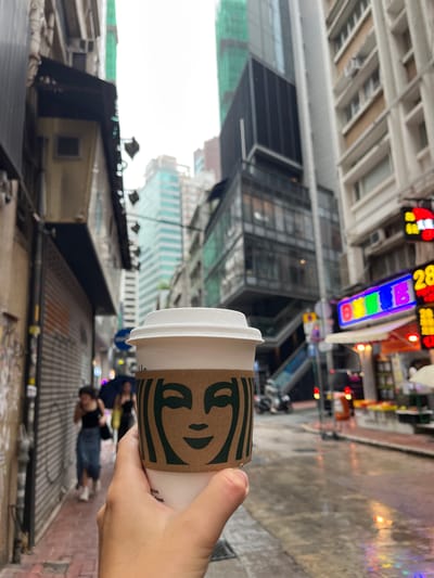 A hand holds a Starbucks coffee cup in front of a blurred urban street scene in Hong Kong, China. Buildings of varying heights line the street, with a mix of architectural styles. The street appears wet, possibly from rain. Pedestrians are visible in the background. Signage and storefronts are discernible in the distance, suggesting a commercial area.