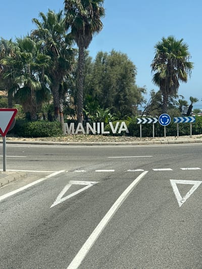 The image depicts a street scene in Manilva, La Chullera, Spain. In the foreground, a road surface is visible with lane markings, including yield triangles painted in white. Behind the road, a large sign with white, three-dimensional letters spells out "MANILVA." Surrounding the sign is landscaping with green bushes and gravel. Several palm trees are prominent in the background, along with other trees and foliage. Road signs are present, including directional chevron signs and a circular blue sign indicating a roundabout. The sky is clear and blue. The overall impression is of a sunny day at a roadside entrance to the town.