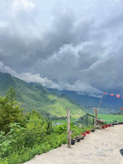 A wide shot captures a dramatic, overcast sky above lush green mountains in Lùng Tám Commune, Vietnam. The foreground features a concrete pathway lined with potted plants, with a rustic wooden post and a small table with benches. A series of red flags with yellow stars are strung on a bamboo pole, extending to the right of the frame. The middle ground reveals a valley with scattered houses and vibrant green fields, nestled between the imposing, mist-shrouded mountains. The overall atmosphere is one of natural grandeur and a sense of remote beauty.