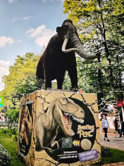 A statue of a mammoth stands on a pedestal decorated with images of dinosaurs. The pedestal has text in Russian that reads "ДиноПарк 'Рекс'" and "КИнь МОНЕТКУ И Я оживУ!" The text indicates the location is Dinopark "Reks" in Stavropol, Russia. In the background, trees and a blue sky with clouds are visible. Several people are walking in the background.
