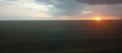 The image depicts a wide, flat, open field under a dramatic sky during what appears to be either sunrise or sunset. The horizon is clearly defined, separating the dark, uniform field from the sky. The sky is a mixture of dark, ominous clouds towards the top and lighter, softer colors towards the horizon, suggesting twilight. A bright, glowing sun is visible near the center-right of the horizon, casting a warm orange and yellow light. Scattered throughout the field, appearing as thin vertical lines, are several utility poles. The overall impression is one of vastness and natural beauty, with a slightly melancholic or serene atmosphere due to the lighting and cloud cover. The location is specified as the city of Adyk, Russia.