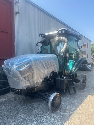 A dark green tractor, partially wrapped in clear protective bubble wrap, is parked on a concrete surface outside a corrugated metal building with a red door on the left. The tractor has a cab with tinted windows and several exterior lights on the roof. The front of the tractor is covered by the bubble wrap, obscuring its details. The tractor's wheels are visible, with the front left wheel removed and lying on the ground. Stacked tires are visible behind the front of the tractor on the left. In the background, the building appears to be a workshop or storage facility. The scene is illuminated by sunlight, casting shadows on the ground. The location is identified as Khryplyn, Ukraine.