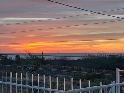 This is a horizontal, eye-level shot of a sunset over a body of water. The sky is filled with horizontal streaks of orange, pink, and purple, with some wispy white clouds. The sun is a bright orange glow near the horizon. In the foreground, there is a white metal fence with vertical posts, some of which are catching the light. Behind the fence is dry, sparse vegetation, and a stone wall. In the mid-ground, there are more dry shrubs and cacti, and beyond that, the body of water. Several thin, dark electrical wires are strung across the sky in the upper portion of the image. The scene appears to be a rural or semi-rural area, possibly along a coastline. The reference to La Guardia, Venezuela, is not discernible from the image itself.