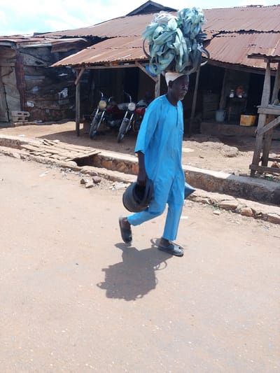 A man walks across a dirt road in Iseyin, Nigeria, carrying a large bundle of light blue and white striped material on his head, balanced by a white cap. He is wearing a long-sleeved blue tunic and trousers. In his right hand, he carries a black bucket. The man's shadow is cast on the ground to his left. In the background, there are several buildings with rusted corrugated iron roofs, appearing to be market stalls or small shops. Two motorcycles are parked in front of one of the structures. The ground is a mix of dirt and gravel, with some scattered debris. The sky is partially visible, with some clouds.