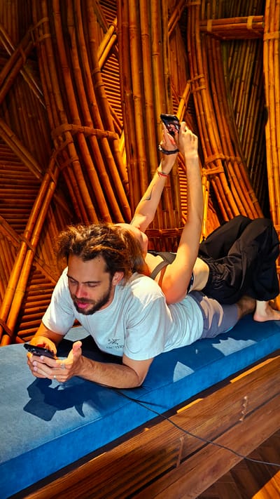 The scene depicts two individuals, a male and a female, situated on a deep blue upholstered bench or daybed, against a backdrop of an intricately constructed bamboo wall, indicative of unique architectural design in Mu Cang Chai, Vietnam.
The male subject lies prone on his stomach, propped on his forearms, with his head slightly raised and turned towards the left of the frame. He has medium-length, dark, curly hair and a full beard. He wears a light grey short-sleeved t-shirt with a dark graphic (partially visible as "ARC'TERYX") and light grey shorts. He holds a black smartphone in both hands, observing its screen with focused attention. A silver ring is visible on his right ring finger.
The female subject is positioned lying across the male's lower back, with her body oriented approximately perpendicular to his. Her head is tilted back, with her face mostly obscured and her light brown hair falling towards the male's right shoulder. She wears a black sports bra and loose black pants. A tattoo is visible on her left upper arm, and she has multiple colorful bracelets on her wrists. Notably, she holds a black smartphone aloft with both feet, pressing its back against her soles and gripping its sides with her toes, looking up at its screen from her supine, inverted position.
Both individuals are independently engrossed in their respective smartphone activities. The background features a complex arrangement of golden-brown bamboo stalks, some bundled and others interwoven in diagonal and horizontal patterns, creating a textured wall. The foreground reveals a section of a polished wooden floor next to the blue cushion. A black cable is visible, running across the blue cushion and extending towards the wooden floor. Strong, directional lighting from an unseen source illuminates the scene, casting distinct shadows on the blue upholstery.