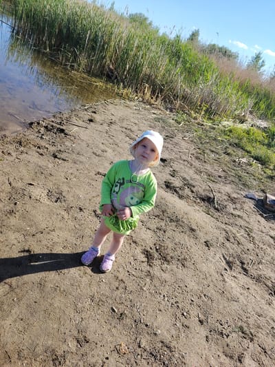 A young child wearing a light-colored bucket hat and a lime green long-sleeved shirt with a graphic print stands on a sandy embankment next to a body of water with tall reeds growing along the edge. The child is wearing light purple sandals with socks. The ground is a mix of sand and dirt, with some footprints visible. The sky is clear and blue with a few wispy clouds. The environment appears to be a natural setting, possibly a lake or riverbank, in the Заямнаўскі сельскі Савет, Belarus region.