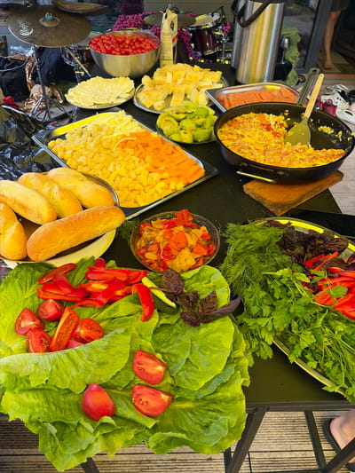 An outdoor food spread is laid out on a dark table, likely in Shebanovo, Russia. In the foreground, a large platter of vibrant green lettuce leaves is topped with halved cherry tomatoes and sliced red bell peppers. Adjacent to this, a tray holds a generous assortment of fresh herbs, including dill and parsley, along with purple basil leaves and more sliced red bell peppers. To the right, a bowl contains cooked vegetables such as peppers and onions. Further back, a pan holds a cooked egg dish, possibly scrambled eggs with bacon, resting on a wooden board. A separate tray displays thin slices of salmon. Other items on the table include several loaves of bread, cubed cheeses in yellow and orange hues, and a bowl filled with small red tomatoes. In the background, parts of a drum set are visible, along with a large stainless steel thermos or beverage dispenser and various bags and fabrics with animal prints. The scene is illuminated by natural light.