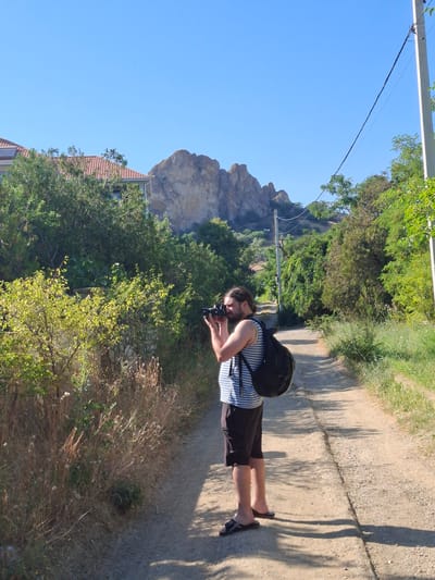 A man with a beard and a backpack is standing on a dirt road, taking a photo with a camera. He is wearing a striped tank top and shorts. The road is lined with trees and bushes, and in the background, there are rocky hills under a clear blue sky. There is a utility pole with wires on the right side of the road. A building with a red tile roof is visible behind the trees on the left. The scene appears to be in a rural or semi-rural area within the Щебетовский поселковый совет, Ukraine, given the context of the request.